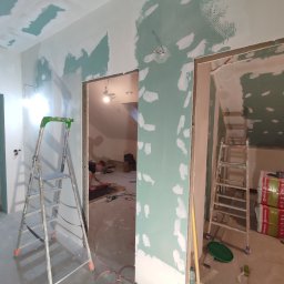 Firma Budowlana SOLARIS - Wnętrze w trakcie prac wykończeniowych: ściany i sufit pokryte wstępną warstwą gładzi, widoczne drabiny, materiały budowlane i narzędzia, w tle osoba pracująca przy podłodze.