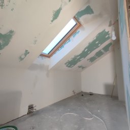 Firma Budowlana SOLARIS - Poddasze w trakcie szpachlowania, widoczne ślady po nałożonej gładzi na skośnym suficie z oknem dachowym, surowy beton na podłodze i wystające rury przy ścianie.