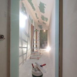 Firma Budowlana SOLARIS - Korytarz w trakcie remontu: ściany z nałożoną gładzią szpachlową, częściowo pokryte płytami karton-gips, wiadro z narzędziami do gładziowania na podłodze, drabina rozstawiona w głębi.
