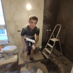 Firma Budowlana SOLARIS - Mężczyzna w trakcie malowania ścian w łazience, widoczny wałek, drabina i częściowo wykończone ściany z imitacją marmuru.