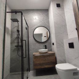 Firma Budowlana SOLARIS - Nowoczesna łazienka z szarymi płytkami imitującymi marmur, kabina prysznicowa z czarnymi profilami, okrągłe lustro nad wiszącą szafką z umywalką oraz podwieszana toaleta.