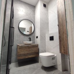 Firma Budowlana SOLARIS - Nowoczesna łazienka z szarymi płytkami imitującymi marmur, wiszącą szafką z umywalką w stylu rustykalnym, okrągłym lustrem w czarnej ramie i podwieszaną toaletą.