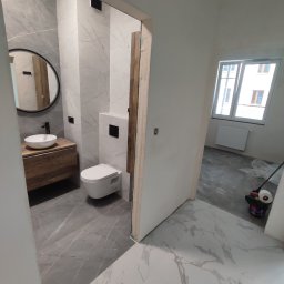 Firma Budowlana SOLARIS - Nowoczesna łazienka z szarymi i białymi marmurkowymi płytkami, drewnianą szafką pod umywalką z okrągłym lustrem i podwieszaną toaletą, widok na sąsiednie pomieszczenie z wiadrem i wałkiem malarskim.