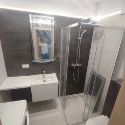 Firma Budowlana SOLARIS - Nowoczesna łazienka w trakcie wykańczania, z brązowymi płytkami na ścianach, białą umywalką z szafką, kabiną prysznicową i lustrem zabezpieczonym folią ochronną, widoczna pralka i toaleta.