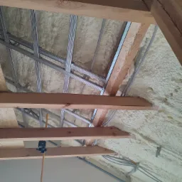 Sufit poddasza z drewnianą konstrukcją, metalowymi profilami i pianką izolacyjną, widoczne kable elektryczne w peszlach i niwelator laserowy na statywie.