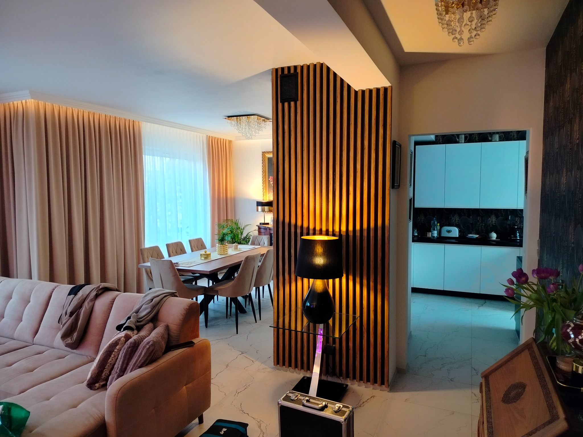 Przestronne wnętrze domu z beżową sofą, drewnianą ścianką działową z oświetleniem, stołem jadalnym i widoczną kuchnią w tle.