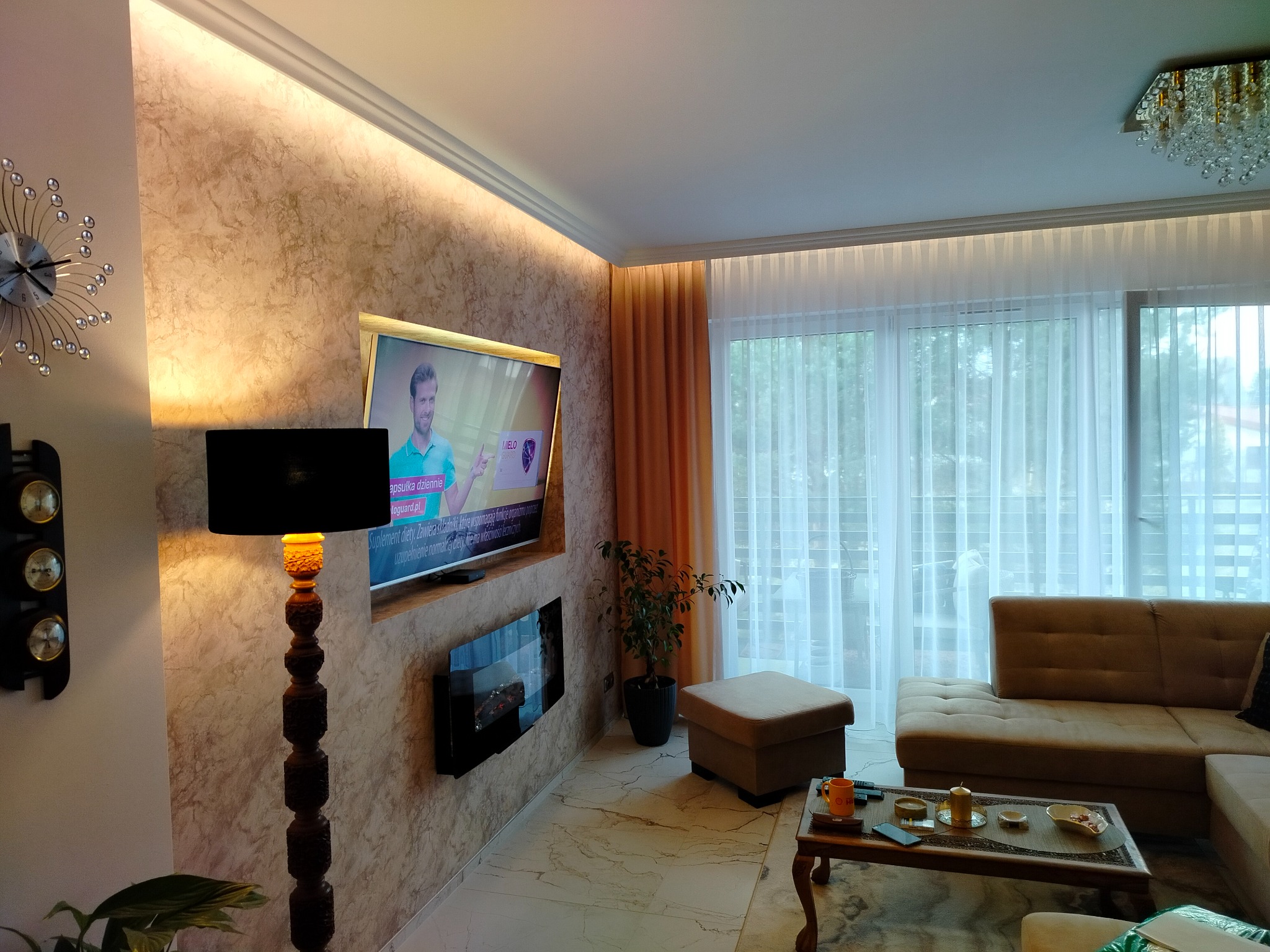 Elegancki salon z dekoracyjną ścianą w odcieniach beżu i złota, podświetloną listwą LED, z telewizorem, kominkiem elektrycznym, drewnianą lampą podłogową i narożną sofą.