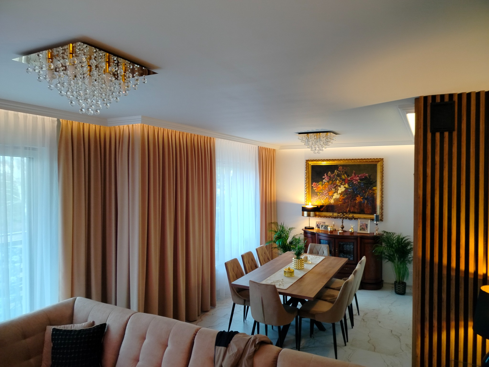Eleganckie wnętrze salonu z jadalnią, beżowe zasłony, kryształowy żyrandol, drewniany stół z krzesłami i obraz w złotej ramie.