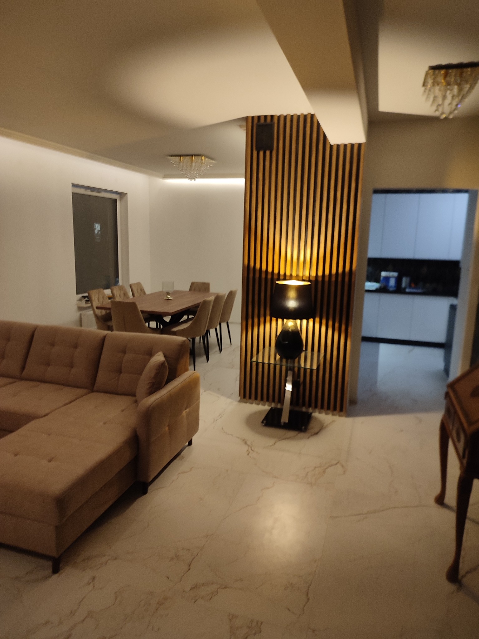 Jasne wnętrze salonu z beżową sofą narożną, marmurową podłogą, drewnianą ścianką działową z oświetleniem oraz stołem jadalnym w tle.