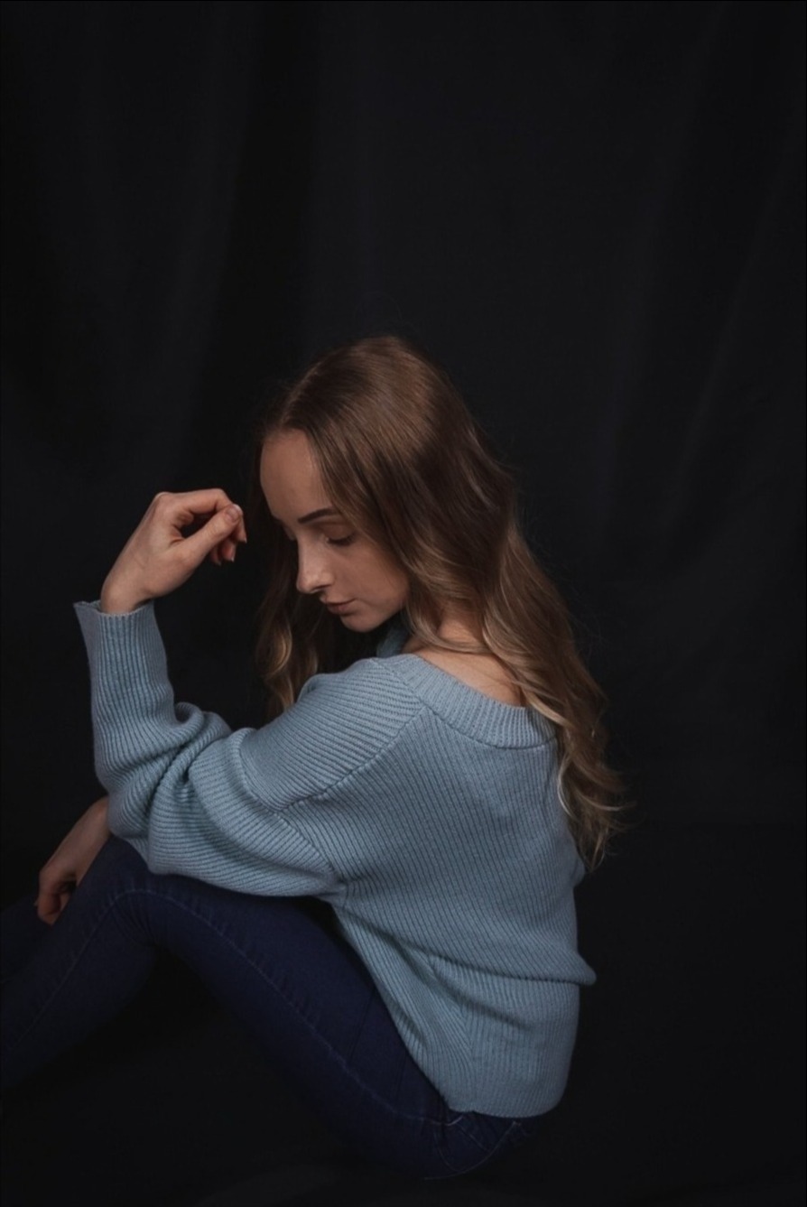 Portret kobiety siedzącej na podłodze, z głową pochyloną w dół i dłonią opartą o czoło, ubrana w błękitny sweter i ciemne jeansy na czarnym tle.