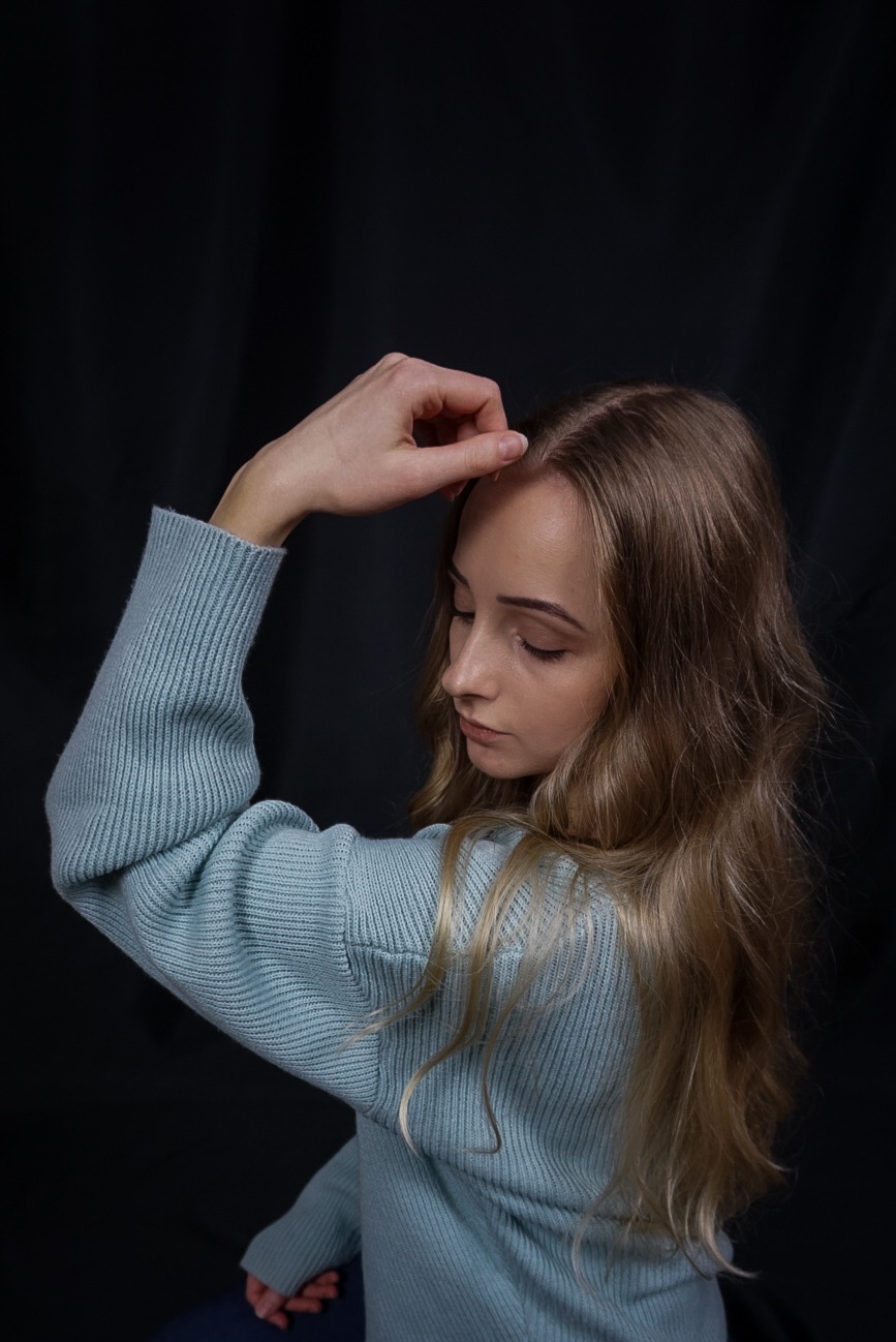 Portret kobiety z długimi blond włosami, ubrana w błękitny sweter, z ręką dotykającą czoła na ciemnym tle.