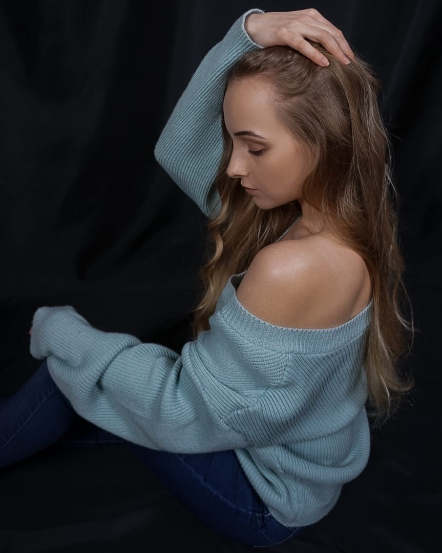 Portret kobiety w Koninie, w niebieskim swetrze z odkrytym ramieniem i jeansach, z dłonią na głowie, na ciemnym tle.