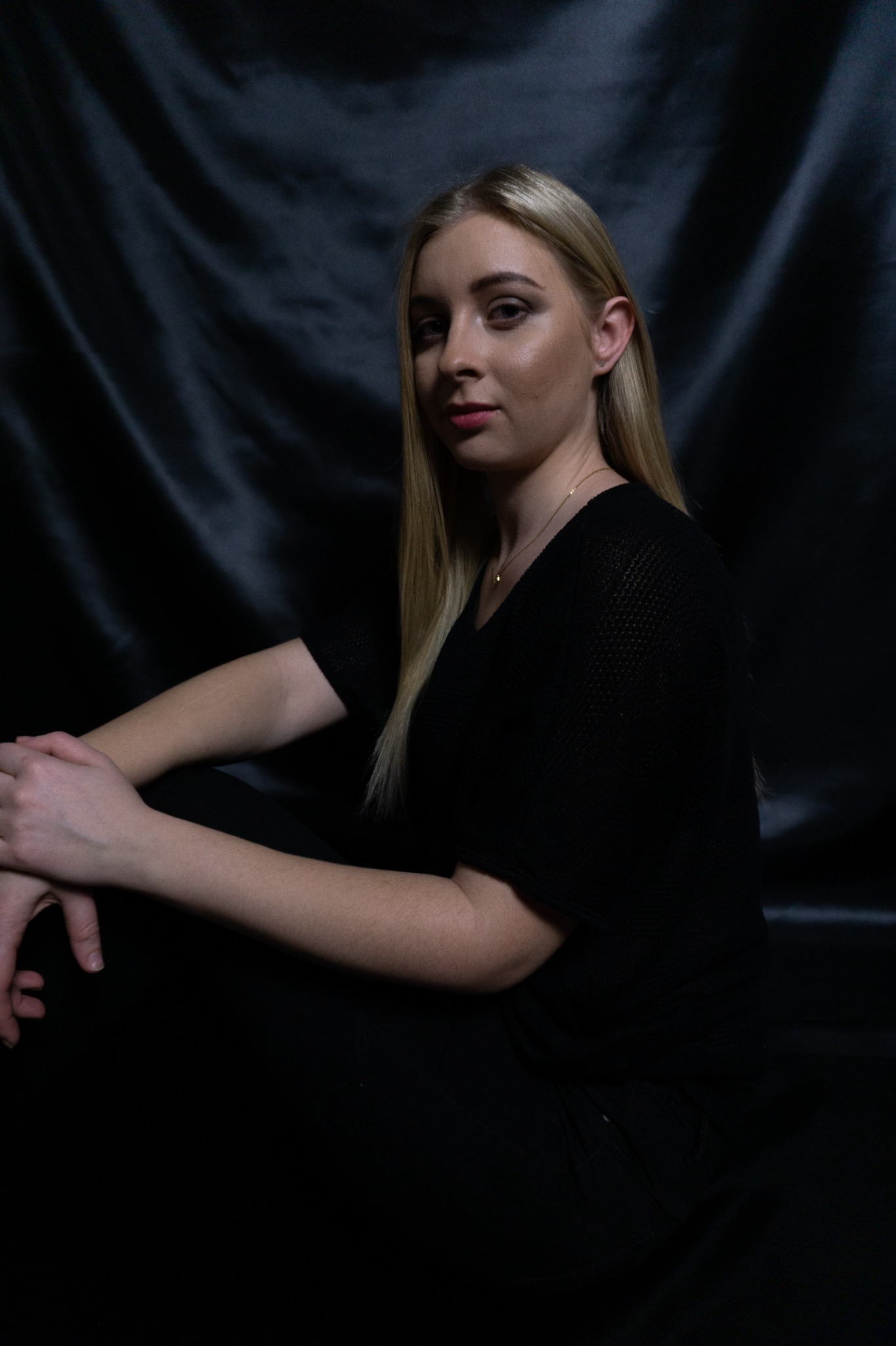Portret młodej kobiety z blond włosami, ubrana na czarno, pozująca na tle czarnej, drapowanej tkaniny.