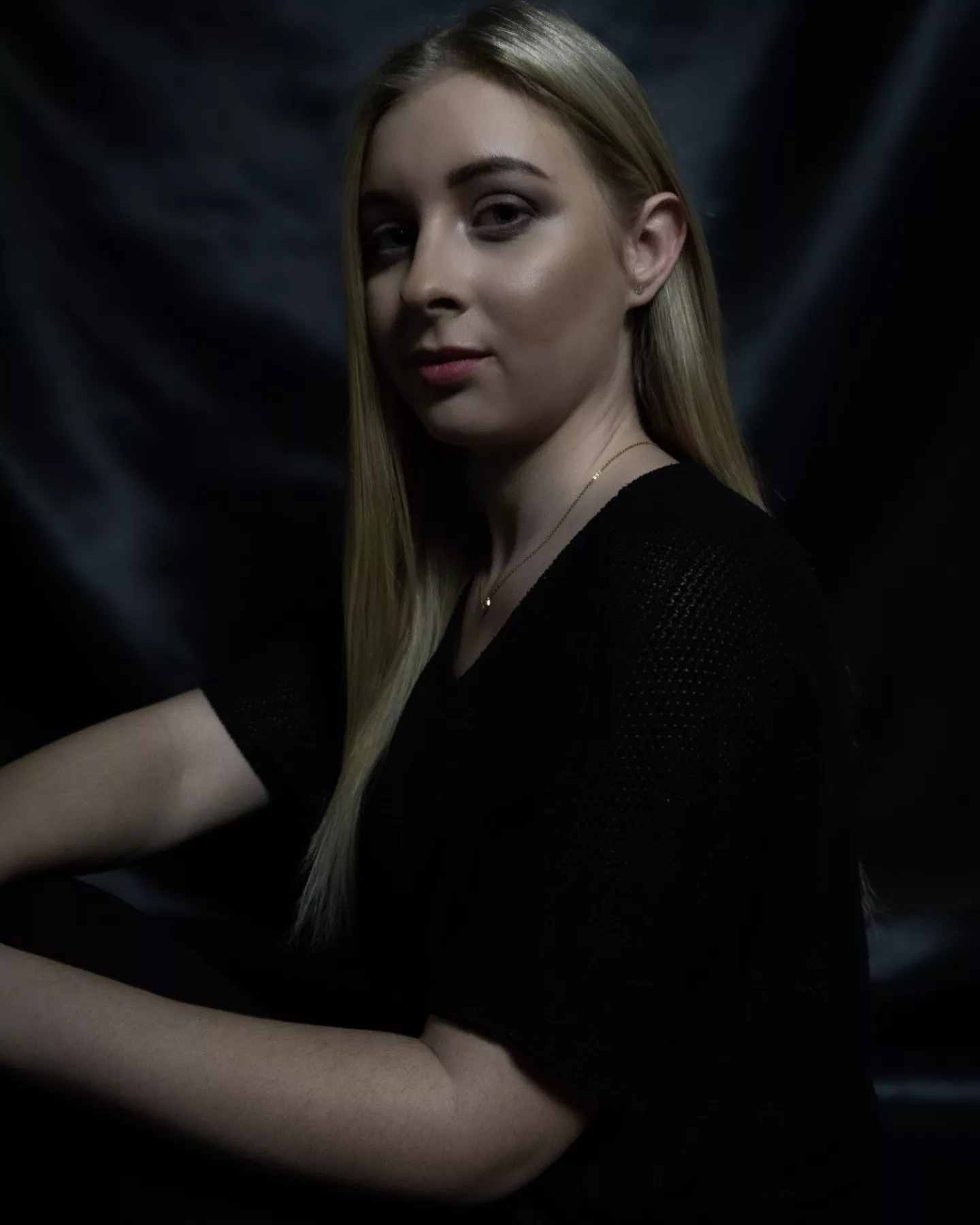 Portret młodej kobiety z blond włosami na ciemnym, drapowanym tle, w czarnym swetrze z delikatnym łańcuszkiem na szyi.