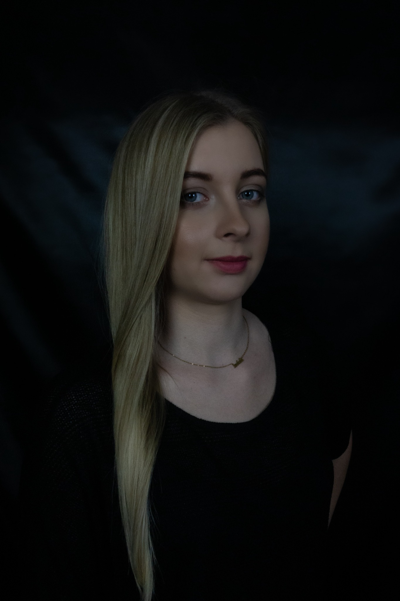 Portret młodej kobiety z długimi blond włosami na ciemnym tle, delikatny uśmiech, złoty naszyjnik z napisem.