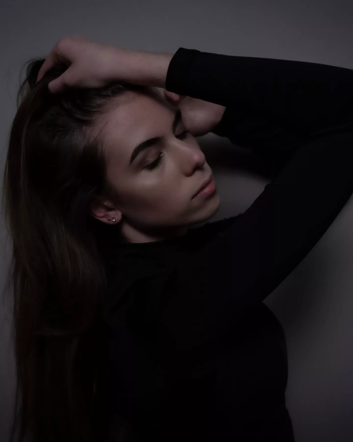 Portret kobiety z długimi brązowymi włosami, w czarnym golfie, z ręką na głowie, pochylona w dół, w ciemnym otoczeniu.