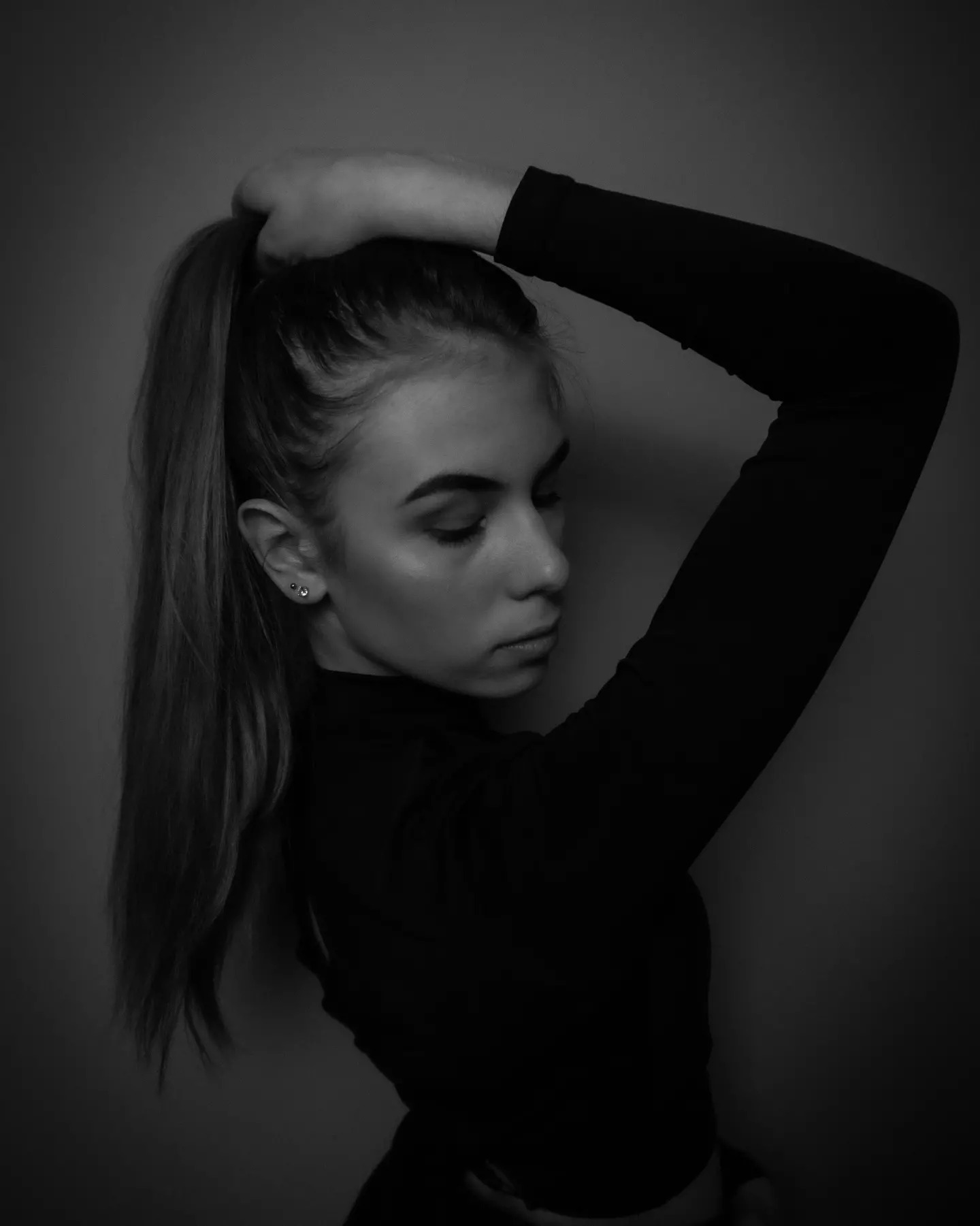 Czarno-biały portret kobiety z podniesioną ręką, upinającej włosy w kucyk, z widocznymi kolczykami w uchu, zamyślony wyraz twarzy.