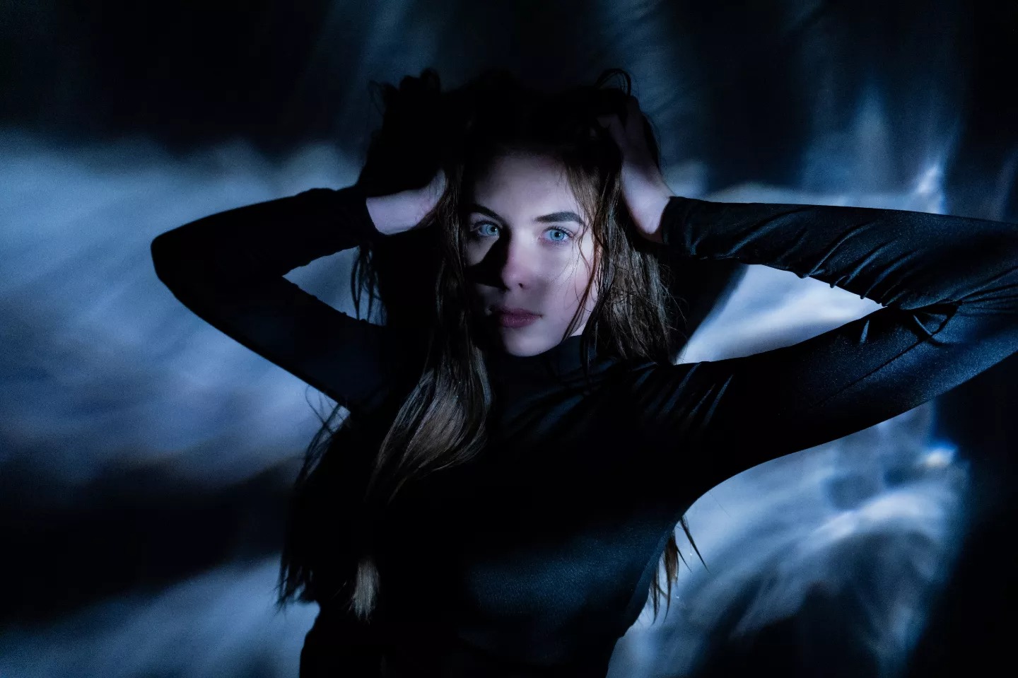 Portret kobiety w czarnym golfie na ciemnym tle z rozproszonym niebieskim światłem, ręce uniesione do włosów, intensywne spojrzenie.