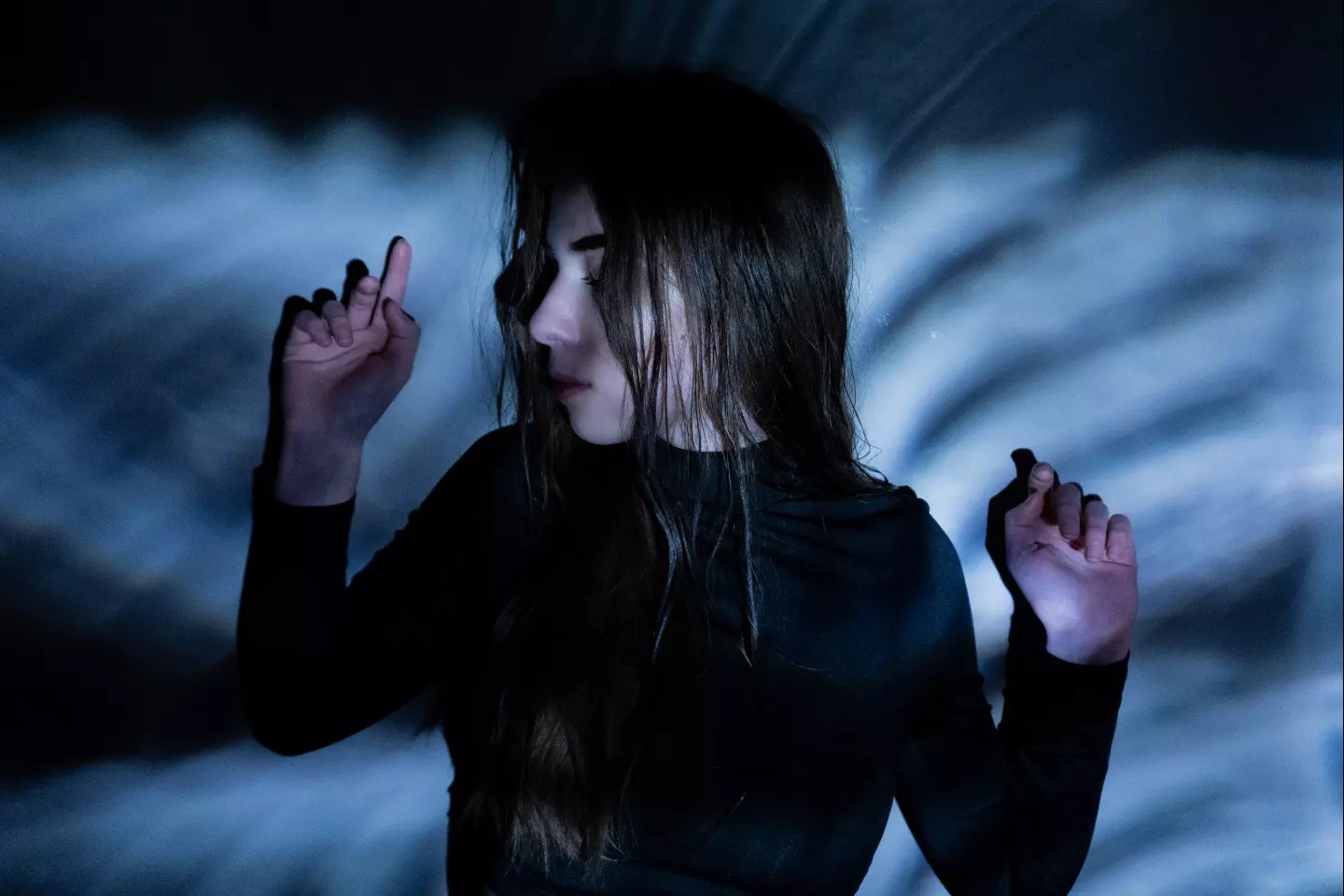 Młoda kobieta w czarnym golfie pozuje na tle abstrakcyjnego, niebieskiego tła z efektem rozmycia, unosząc dłonie.