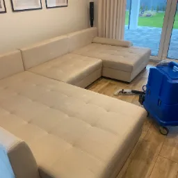 Jasnobeżowa, modułowa kanapa narożna z pikowanymi siedziskami, obok której stoi niebieski odkurzacz piorący na drewnianej podłodze w salonie z widokiem na ogród.