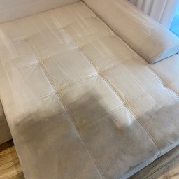 Zatoka Błysku Robert Mastela - Kremowa kanapa z widoczną różnicą w czystości tapicerki po praniu, z poduszką wałkiem. Część kanapy po lewej stronie jest czystsza od prawej.