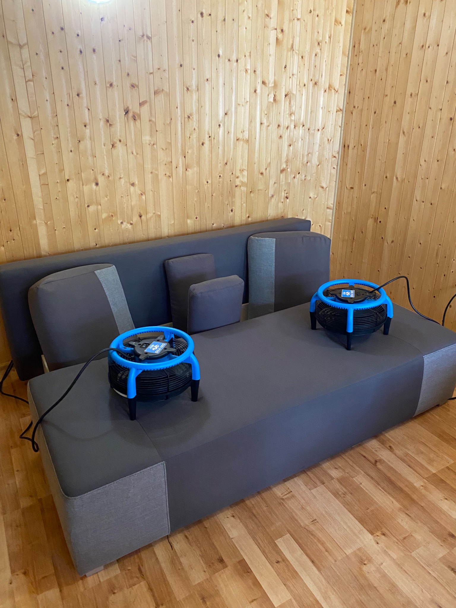 Szara kanapa z dwoma niebieskimi wentylatorami do suszenia tapicerki, ustawionymi na siedzisku. Drewniana ściana w tle.
