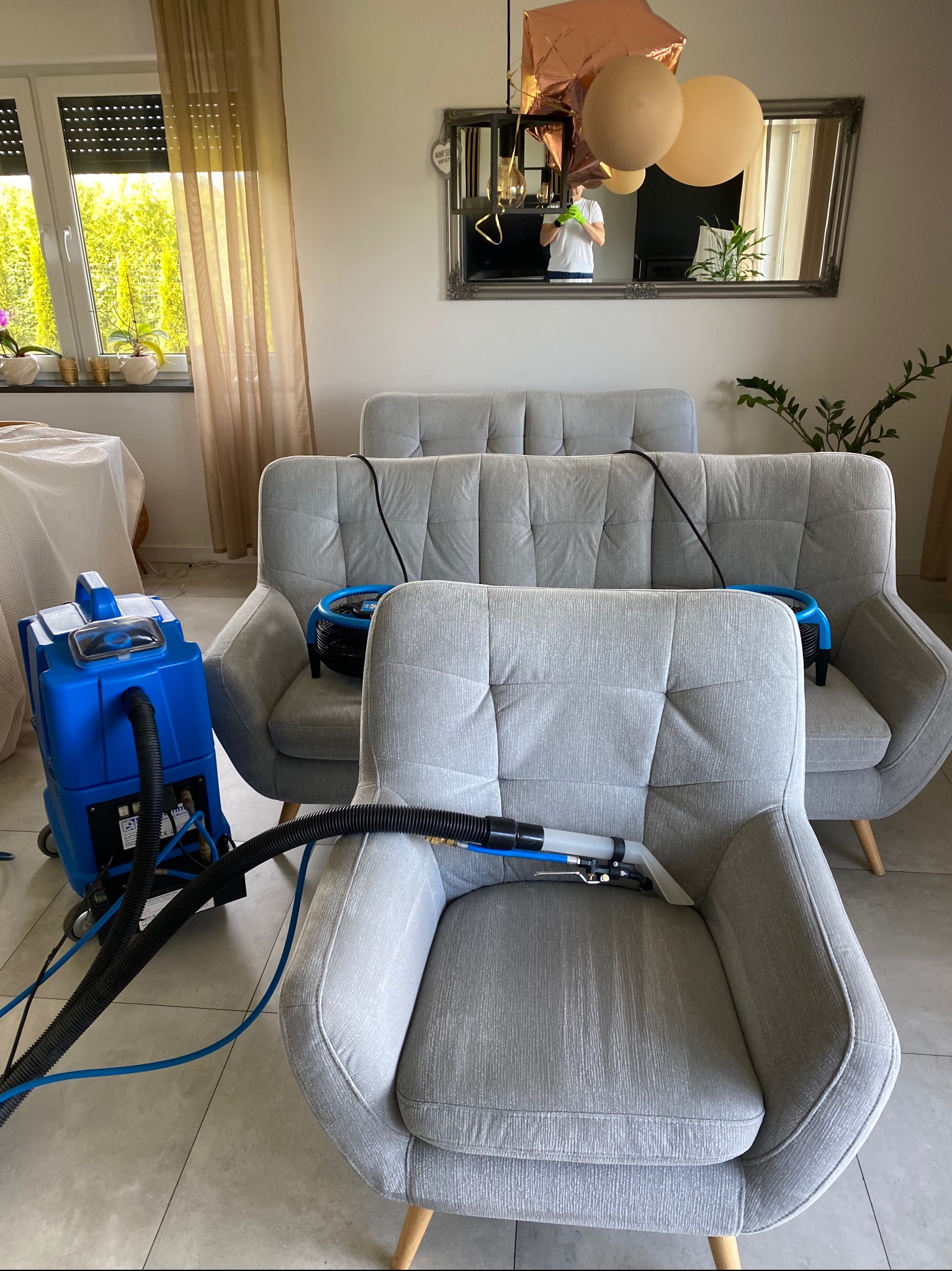 Fotel i sofa w trakcie czyszczenia parowego, widoczny sprzęt czyszczący, w tle okno i lustro z odbiciem osoby wykonującej usługę.