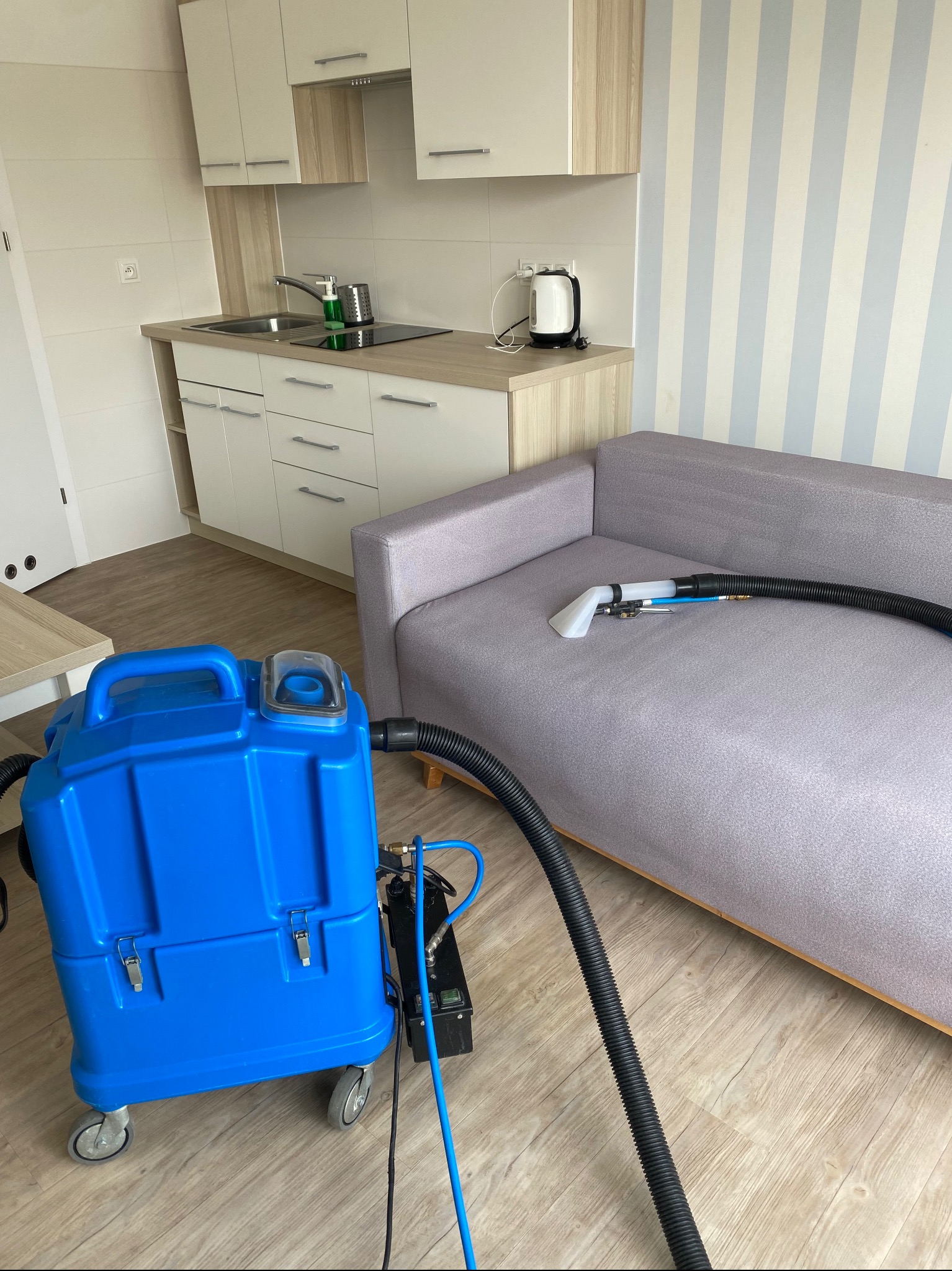 Profesjonalny sprzęt do prania tapicerki w trakcie czyszczenia jasnoszarej sofy w mieszkaniu z aneksem kuchennym i ścianą w biało-niebieskie pasy.