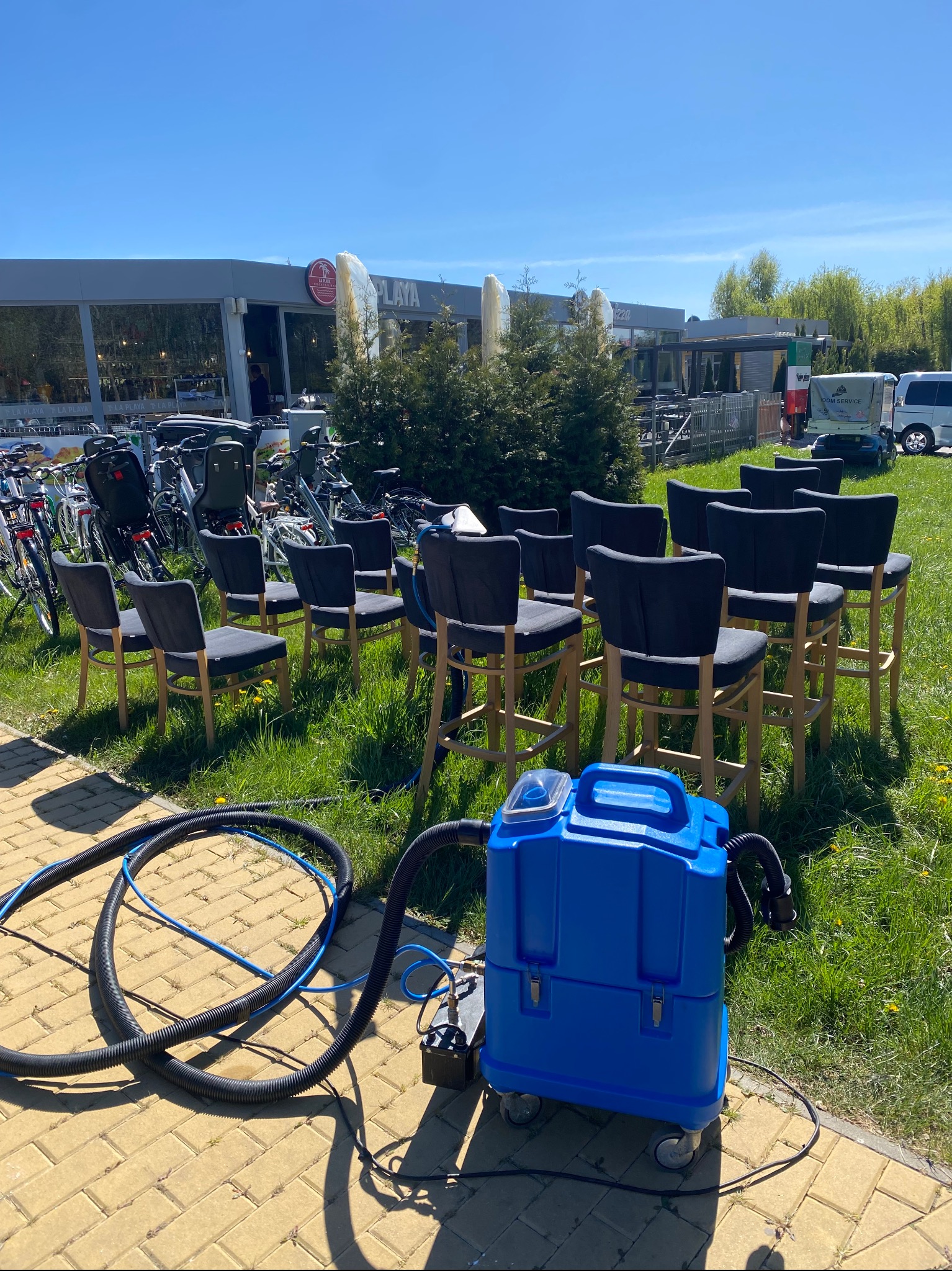 Niebieski odkurzacz piorący na trawie przed rzędem krzeseł barowych z czarną tapicerką. W tle budynek La Playa i zaparkowane rowery.