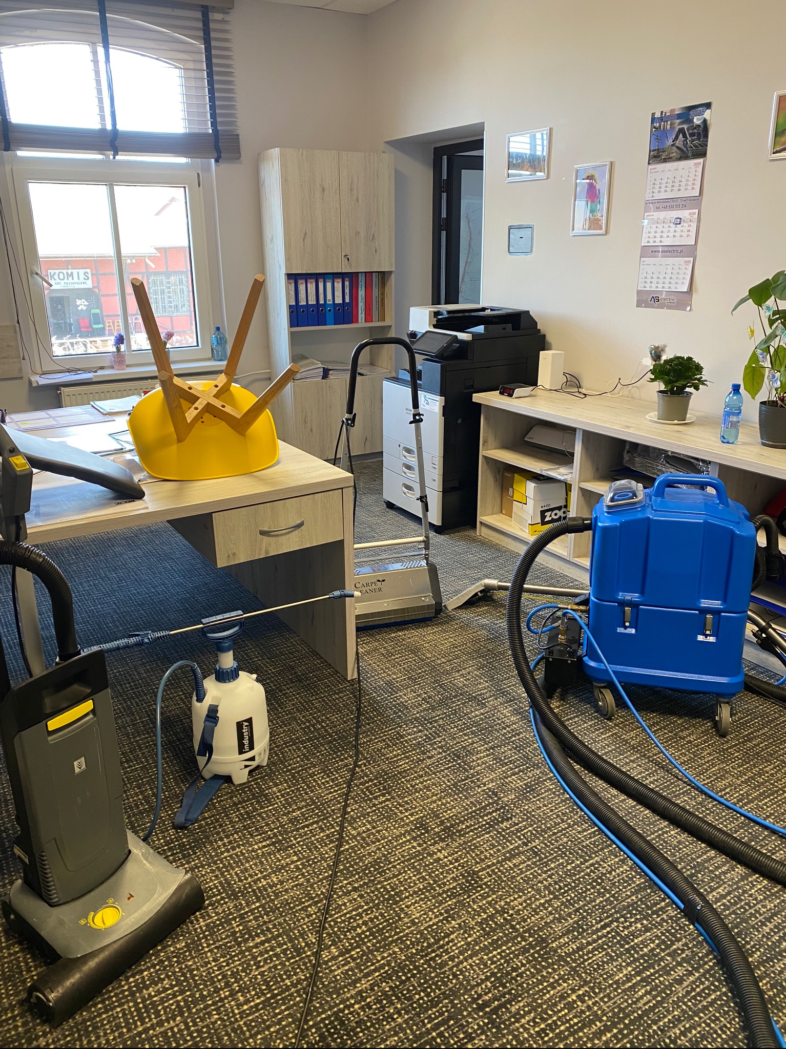 Sprzęt do czyszczenia dywanów w biurze: odkurzacz piorący, niebieski zbiornik na kółkach, przewody. Żółte krzesło odwrócone na biurku. Dywan z wzorem.