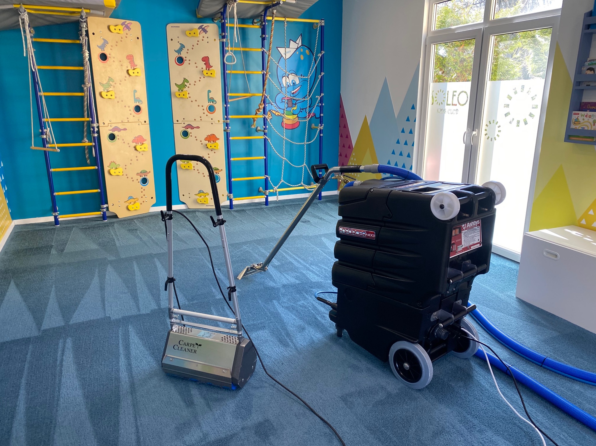Profesjonalne czyszczenie dywanu w pokoju zabaw dla dzieci z użyciem specjalistycznego sprzętu. Widoczne ślady po czyszczeniu na niebieskim dywanie.
