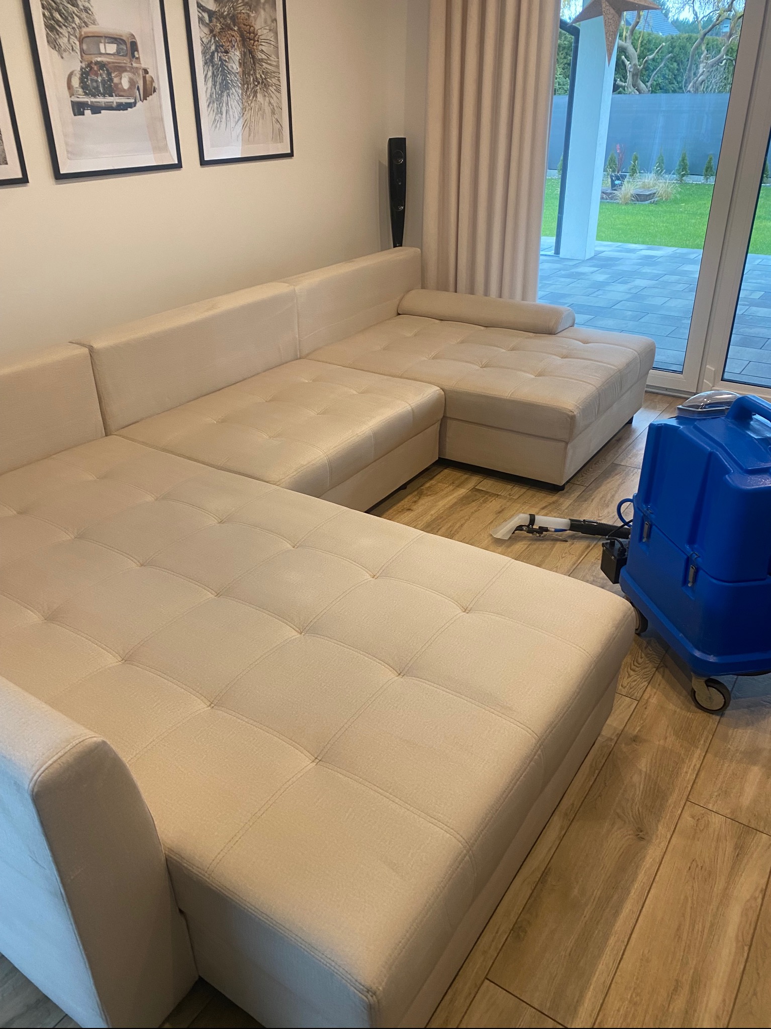 Jasnobeżowa, modułowa kanapa narożna z pikowanymi siedziskami, obok której stoi niebieski odkurzacz piorący na drewnianej podłodze w salonie z widokiem na ogród.