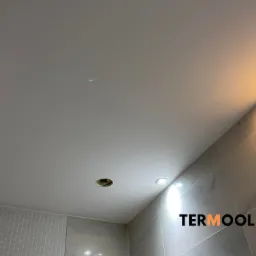 Biały sufit podwieszany z zamontowanymi punktowymi lampami LED i otworem na instalację, widoczne płytki ceramiczne na ścianie.