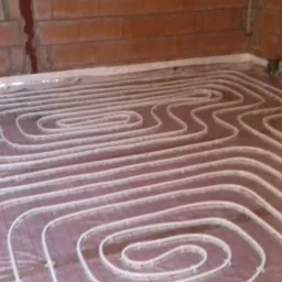 Rozłożone rury ogrzewania podłogowego na folii izolacyjnej w surowym wnętrzu z ceglanymi ścianami, widok z g&oacute;ry pod kątem.