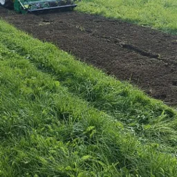 Traktor z glebogryzarką przygotowuje ziemię pod trawnik na zewnątrz, widoczni dwaj mężczyźni w odzieży roboczej.