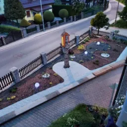 Widok z góry na świeżo zaprojektowany ogród z oświetleniem punktowym, ścieżkami z białego żwiru i centralnie umieszczoną drewnianą kapliczką, otoczony zadbanym trawnikiem i elementami małej...