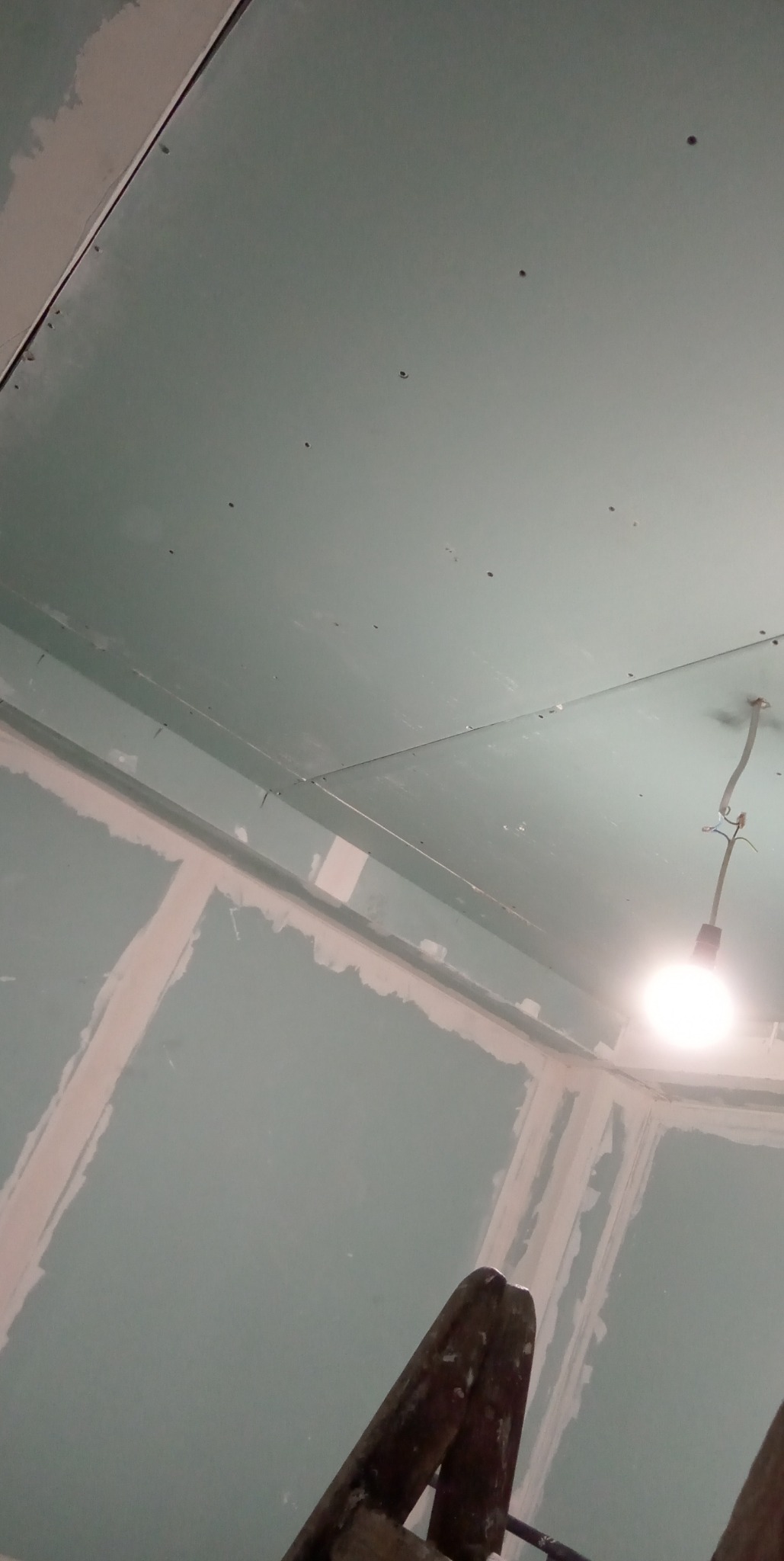 Ściana i sufit w trakcie przygotowania do malowania, widoczne płyty gipsowo-kartonowe, szpachlowane łączenia i wkręty, ujęcie z dołu ze schodów.