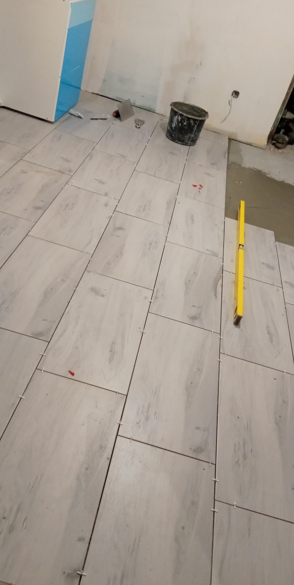 Układanie jasnych, kwadratowych płytek podłogowych z użyciem krzyżyków dystansowych, poziomica w kadrze, widoczne narzędzia i wiadro z zaprawą.