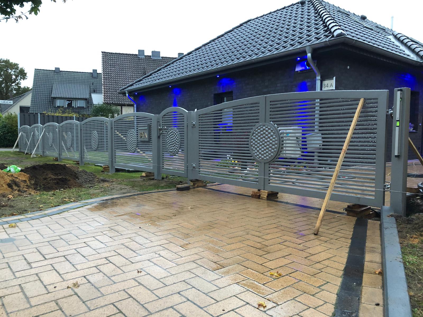 Szara, przesuwna brama wjazdowa z poziomymi szczeblami i okrągłymi dekoracjami na tle domu z niebieskim oświetleniem, widoczne drewniane podpory pod bramą i mokra kostka brukowa.