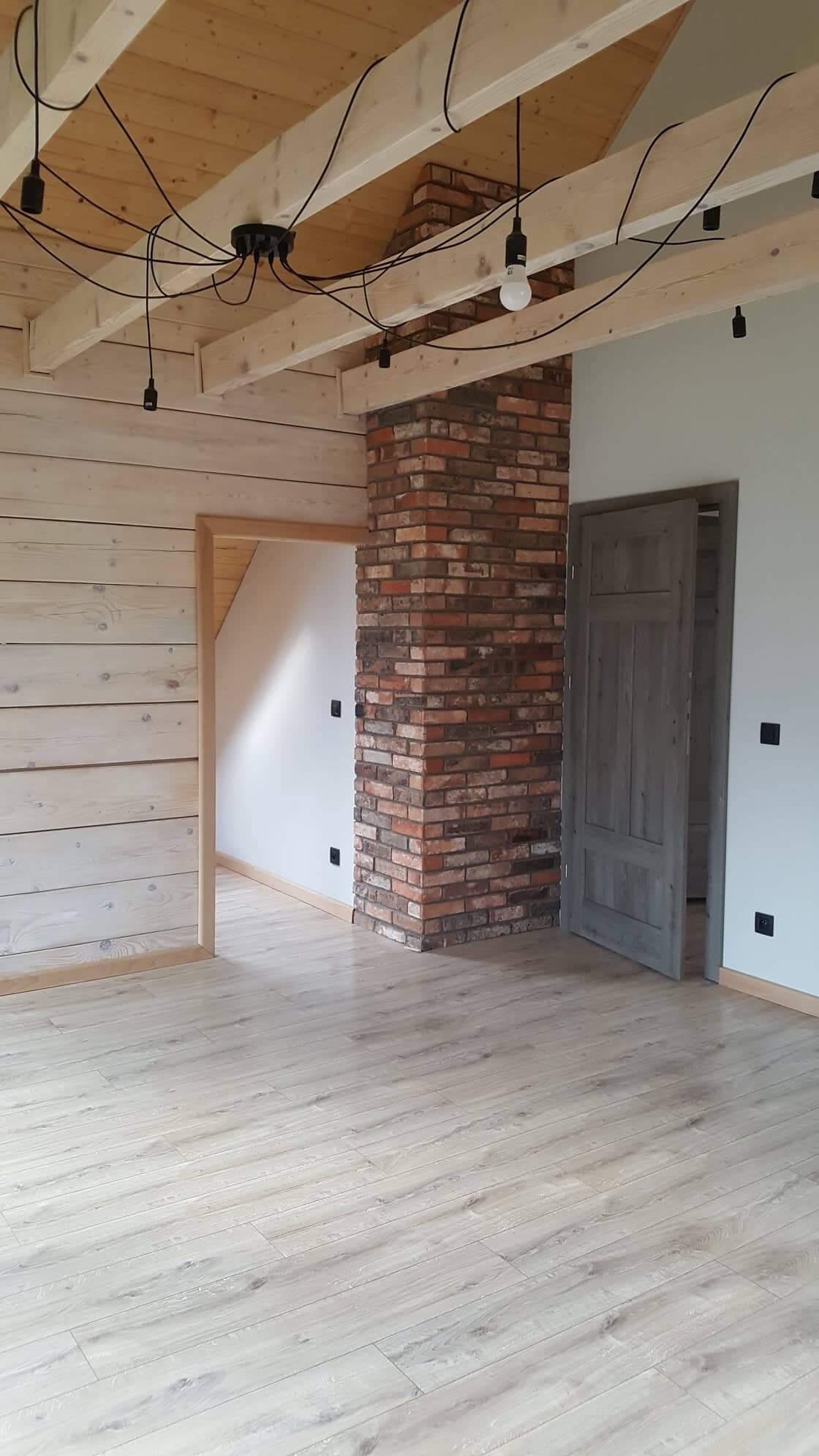Surowe wnętrze z drewnianymi belkami stropowymi, ścianą z desek pomalowanych na biało, kominem z czerwonej cegły i jasną podłogą imitującą drewno.