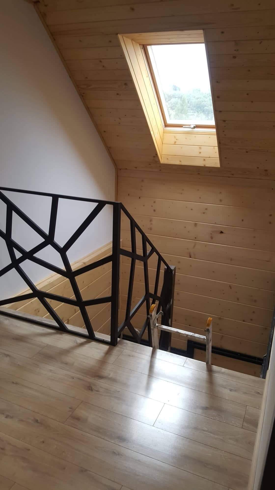 Drewniane panele ścienne i sufitowe na poddaszu z oknem dachowym, czarna, geometryczna balustrada schodów, jasne panele podłogowe i drabina malarska.
