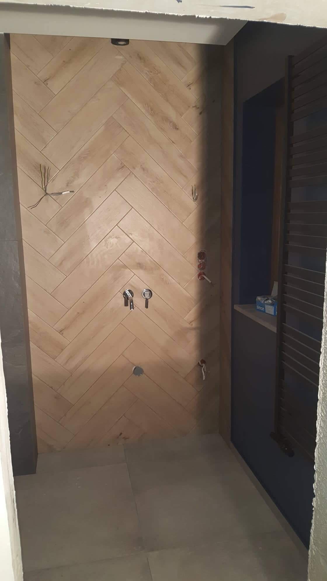 Ściana prysznica wyłożona jasnymi płytkami imitującymi drewno w jodełkę, widoczne przygotowane instalacje wodne i elektryczne, grzejnik drabinkowy na ścianie bocznej, etap prac wykończeniowych.