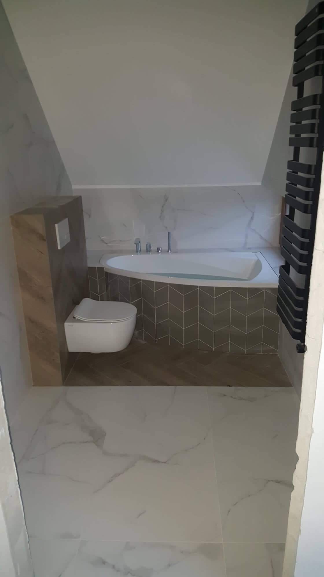 Wykończona łazienka na poddaszu z owalną wanną, wiszącą toaletą i grzejnikiem drabinkowym w kolorze czarnym; ściany wyłożone białymi płytkami imitującymi marmur.