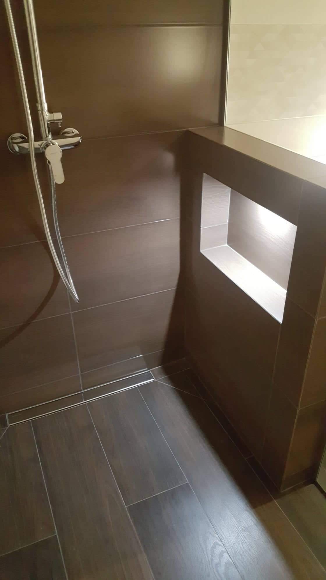 Nowoczesna kabina prysznicowa z ciemnobrązowymi płytkami imitującymi drewno, chromowaną armaturą i odpływem liniowym, z wnęką w ścianie wyłożoną jasnymi płytkami.