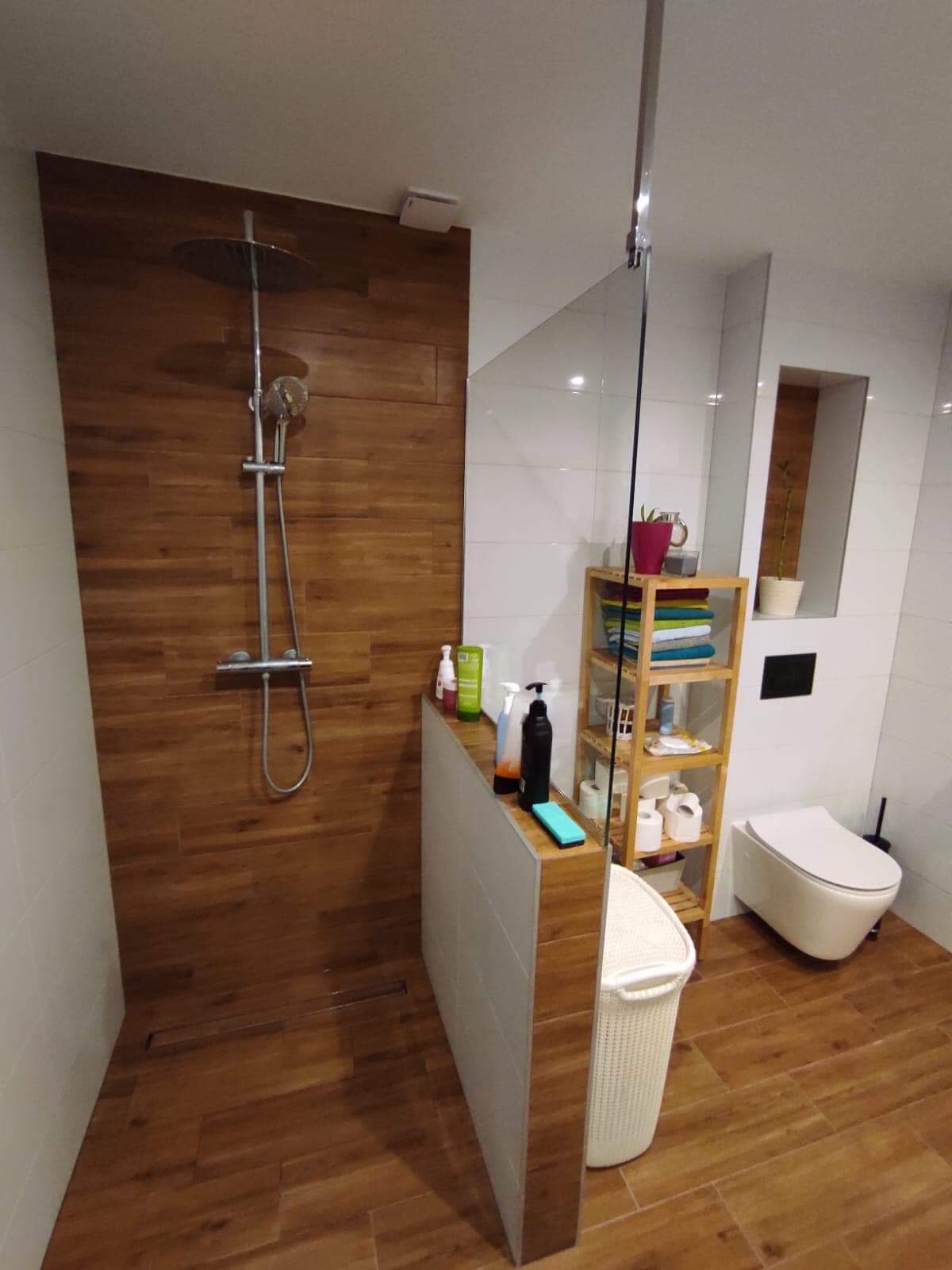 Nowoczesna łazienka z drewnopodobnymi płytkami na podłodze i pod prysznicem, szklana kabina prysznicowa, biała toaleta podwieszana i drewniany regał z ręcznikami oraz kosmetykami.