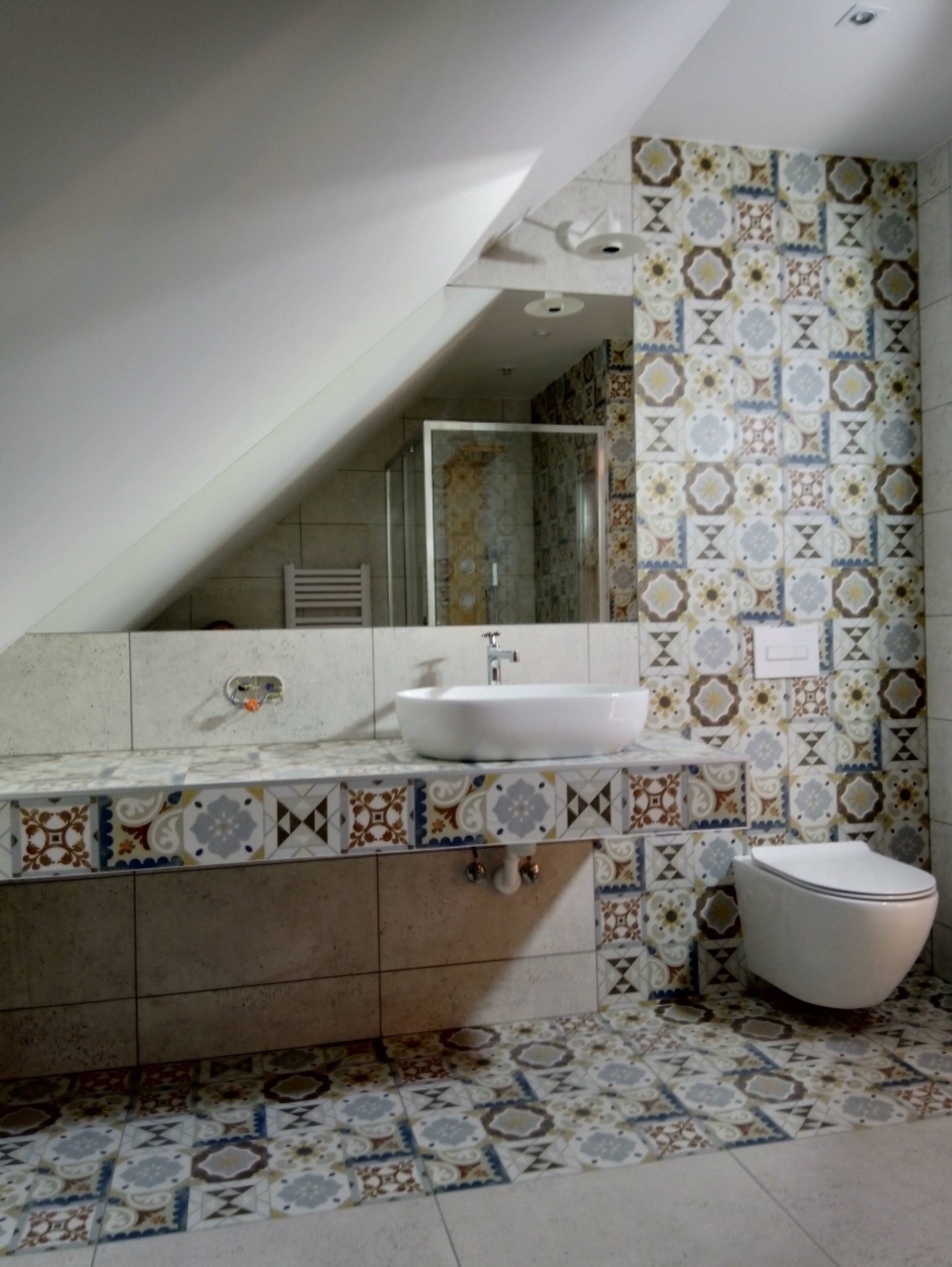 Wykończona łazienka na poddaszu z umywalką nablatową, wiszącą toaletą i wzorzystymi płytkami na ścianach i podłodze, odbicie kabiny prysznicowej w lustrze.