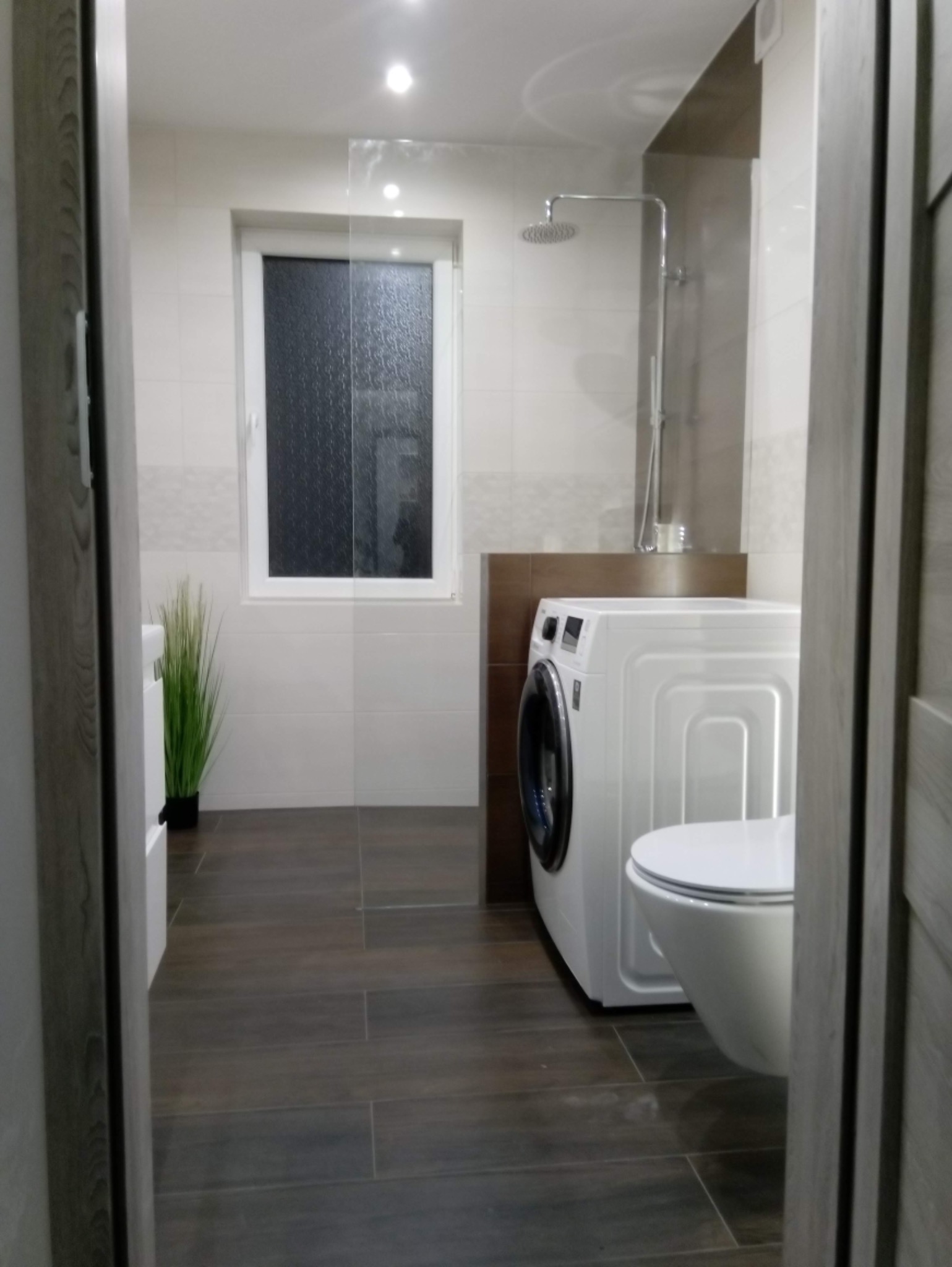 Nowoczesna łazienka z brązowymi płytkami podłogowymi, białą pralką i podwieszaną toaletą, widok zza uchylonych drzwi.