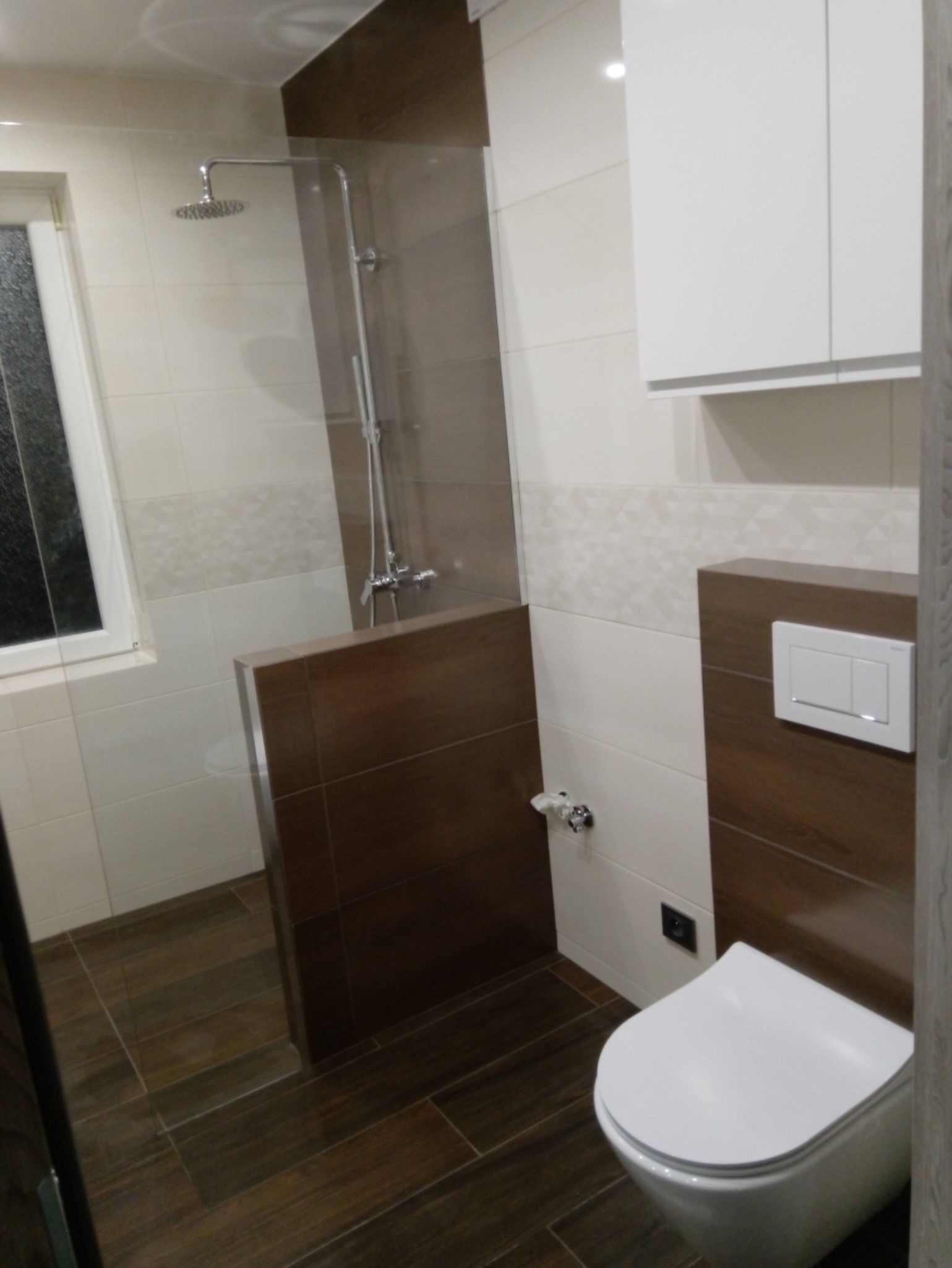 Nowoczesna łazienka z brązowymi i białymi płytkami, kabina prysznicowa typu walk-in z deszczownicą, podwieszana toaleta i biała szafka wisząca.