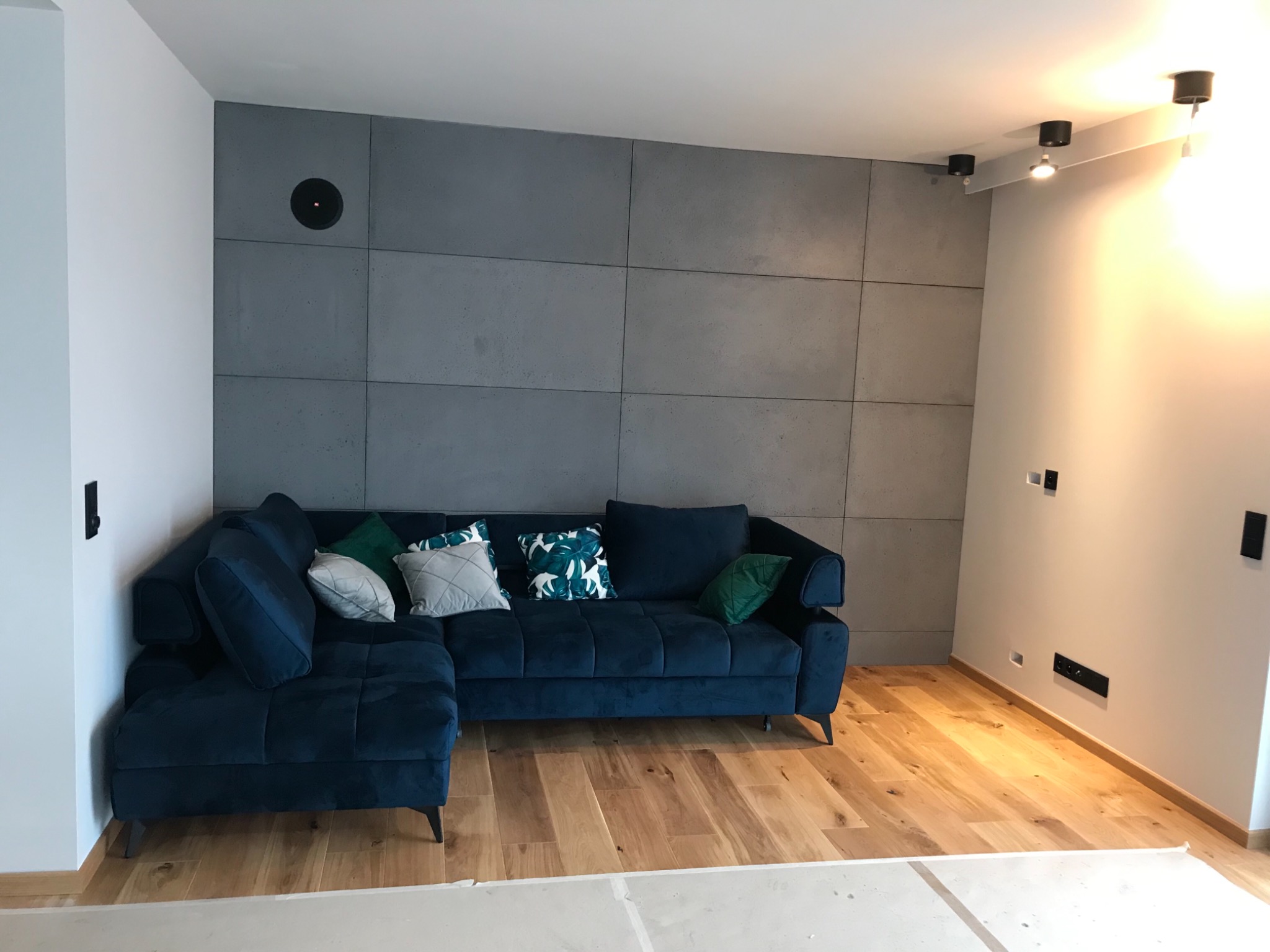 Salon z drewnianą podłogą i ścianą wykończoną betonowymi panelami, oświetleniem punktowym i narożną, granatową sofą z dekoracyjnymi poduszkami.