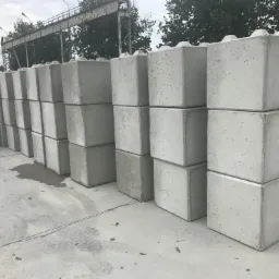 Równy rząd szarych, betonowych bloków prefabrykowanych ułożonych na betonowym placu, z elementami przypominającymi klocki Lego na górze, z widocznymi śladami opon w tle.